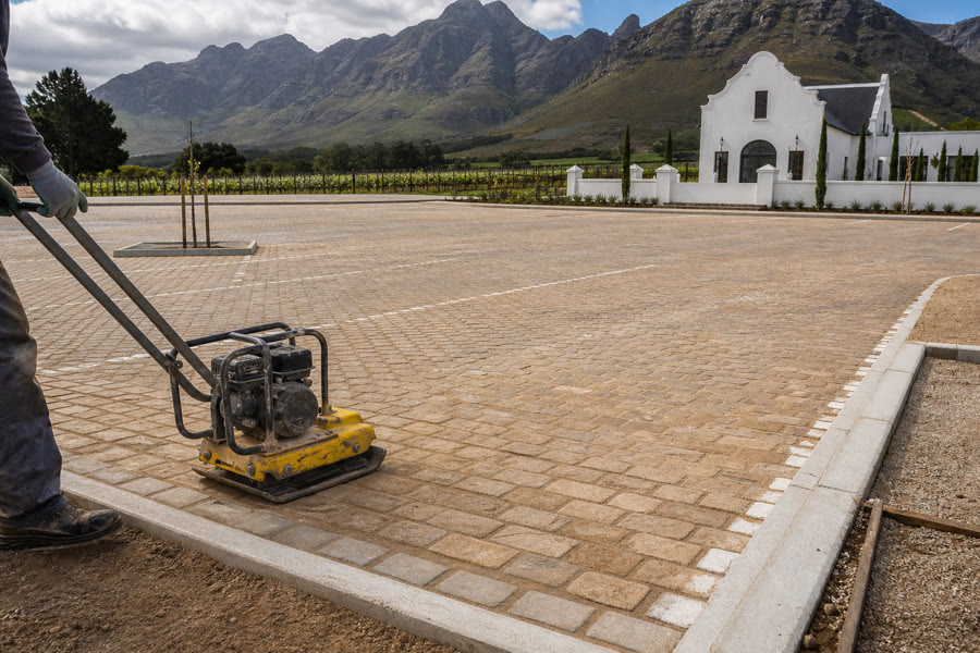 Sandstone cobble commercial parking apron at a Stellenbosch Cape Dutch wine estate, plate compactor on the finished section.